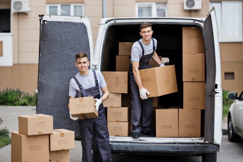 Two young handsome smiling movers wearing uniforms are unloading the van full of boxes. House move